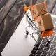 EUA - Imagem de uma miniatura de carrinho de supermercado com duas caixas de papelão dentro, em cima de um teclado de notebook - e-commerce